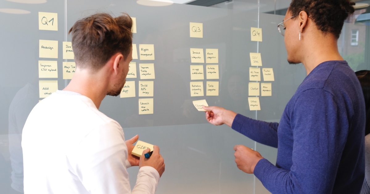 Two men standing at a glass wall collaborating over an agile task list with sticky notes.